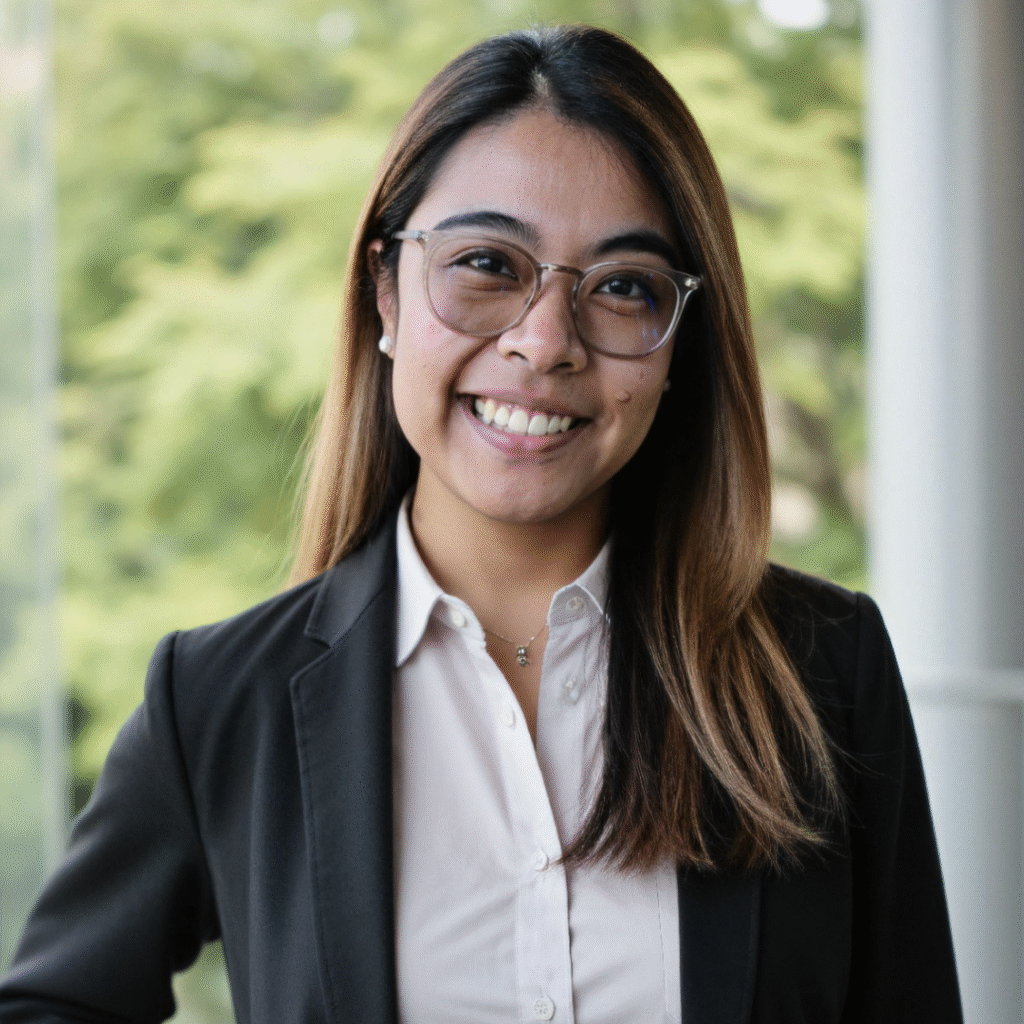 Professional woman with long straight hair wearing glasses, a white blouse, and a black blazer, smiling confidently in front of a blurred green outdoor background.