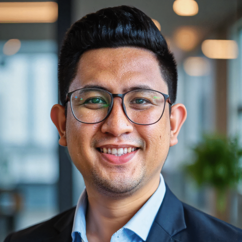 Professional man with short styled hair and glasses, wearing a dark blazer over a light blue shirt, smiling confidently in a modern office setting with blurred lights and greenery in the background.